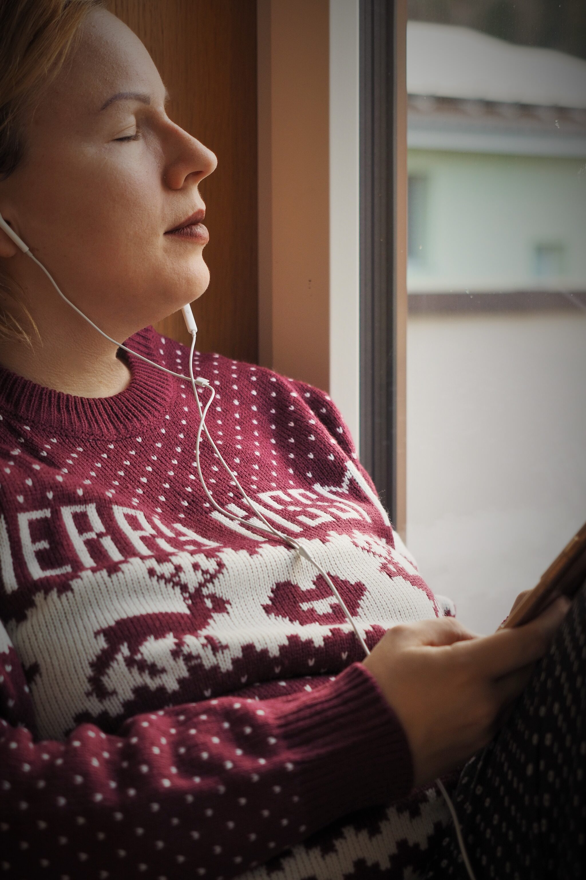 Frau sitzt am Fenster und hört mit geschlossenen Augen Musik über Kopfhörer auf dem Smartphone